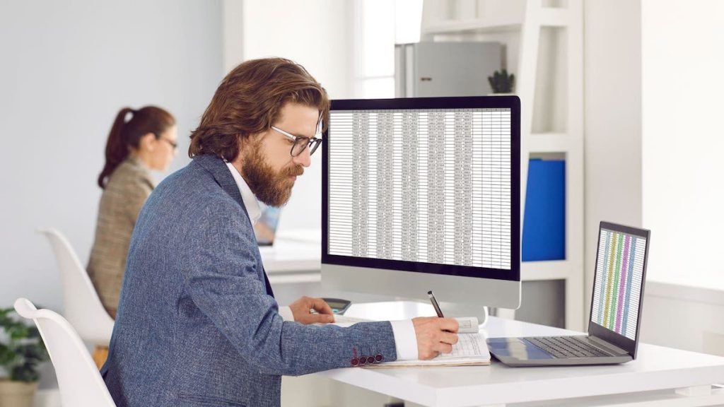 Data manager writing in notebook as report displays on desktop computer | Linkpoint360