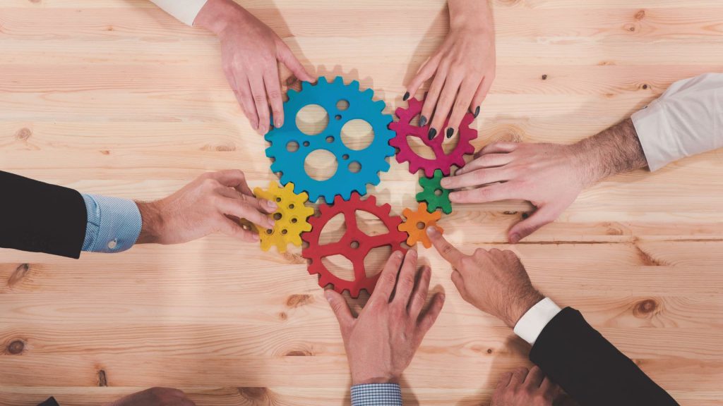 Aerial View of Sales Team Members Grabbing Colorful Gears From Wooden Desk | What is CRM Integration?